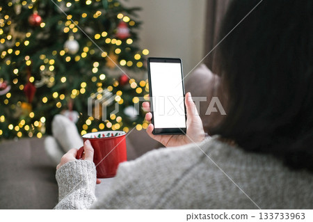Woman in sweater holding red mug and smartphone with blank white screen with blurred Christmas tree and bokeh lights in background. Mobile app, cozy holidays, screen mockup, holiday shopping Woman in sweater holding red mug and smartphone with blank white screen with blurred Christmas tree and bokeh lights in background. Mobile app, cozy holidays, screen mockup, holiday shopping 133733963
