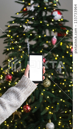 Person in cozy sweater holds smartphone with blank white screen mock-up against backdrop of decorated Christmas tree. Holiday apps, seasonal shopping, digital greetings Person in cozy sweater holds smartphone with blank white screen mock-up against backdrop of decorated Christmas tree. Holiday apps, seasonal shopping, digital greetings 133733964
