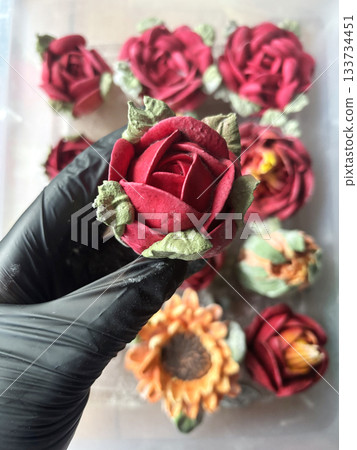 Hand in a rubber glove holding a marshmallow rose, Homemade marshmallow, Marshmallow flowers, Marshmallow roses, tulips and sunflowers are arranged in the background 133734451