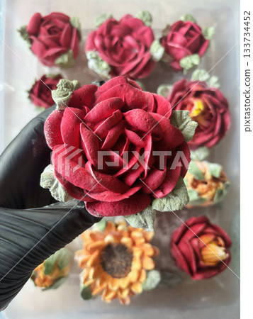 Hand in a rubber glove holding a marshmallow rose, Homemade marshmallow, Marshmallow flowers, Marshmallow roses, tulips and sunflowers are arranged in the background 133734452