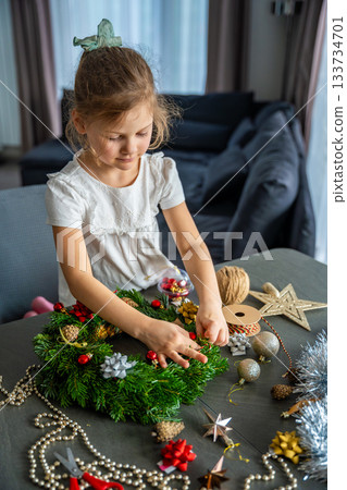 Little girl finishing her handmade Christmas wreath by tying a string to hang it. Final step of a festive DIY project captured at home in a cozy family setting. Little girl finishing her handmade Christmas wreath by tying a string to hang it. Final step of a festive DIY project captured at home in a cozy family setting. 133734701