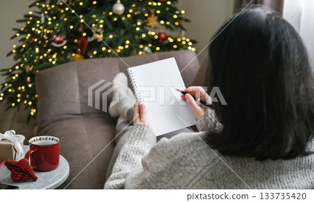 Woman in cozy sweater writing in blank spiral notebook while relaxing on sofa with festive Christmas tree in background. Planning, resolutions, to-do list, wish list, mockup, shopping list Woman in cozy sweater writing in blank spiral notebook while relaxing on sofa with festive Christmas tree in background. Planning, resolutions, to-do list, wish list, mockup, shopping list 133735420