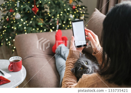 Woman in cozy sweater and red socks holding smartphone with blank white screen relaxing on sofa with cat during Christmas holidays. Holiday apps, mobile shopping, seasonal social media Woman in cozy sweater and red socks holding smartphone with blank white screen relaxing on sofa with cat during Christmas holidays. Holiday apps, mobile shopping, seasonal social media 133735421