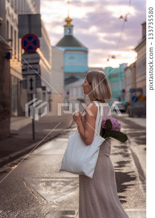 A woman stands on a wet city street at sunset with a white canvas tote and pink hydrangea, framed by soft pastel tones and historic buildings A woman stands on a wet city street at sunset with a white canvas tote and pink hydrangea, framed by soft pastel tones and historic buildings 133735580