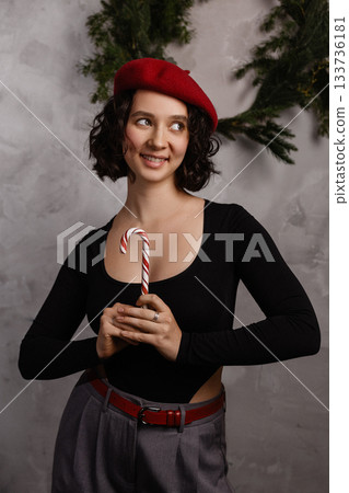 Caucasian woman holding candy cane in festive room with grey wall and green garland Caucasian woman holding candy cane in festive room with grey wall and green garland 133736181