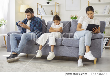 Sad child ignored as parents use devices on couch at home, family disconnect Sad child ignored as parents use devices on couch at home, family disconnect 133736635