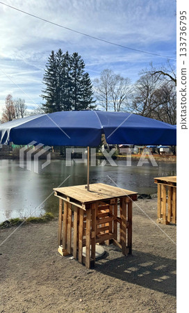Cozy Outdoor Bar Tables Under a Blue Umbrella By a Serene Pond Setting for Relaxed Weekend Leisure Cozy Outdoor Bar Tables Under a Blue Umbrella By a Serene Pond Setting for Relaxed Weekend Leisure 133736795
