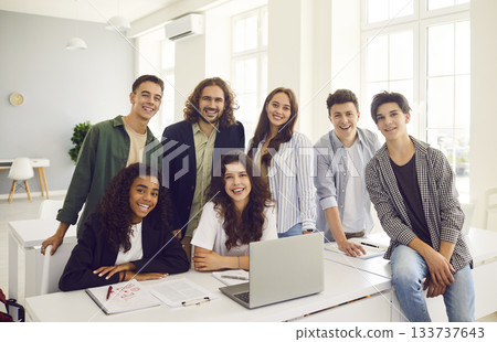 Classmates sitting in classroom with male friendly teacher at the desk and looking at camera. 133737643