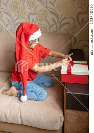 A girl in a Santa Claus hat opens a gift. It's Christmas time. A girl in a Santa Claus hat opens a gift. It's Christmas time. 133738150