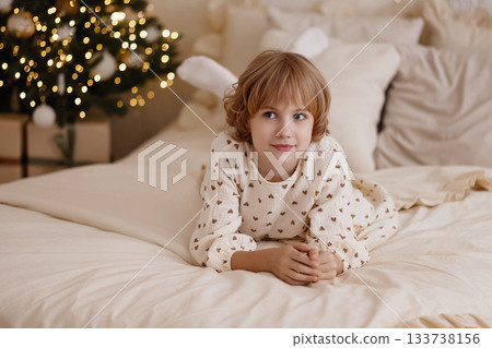 Caucasian child lying on bed in cozy bedroom with Christmas tree, look at camera Caucasian child lying on bed in cozy bedroom with Christmas tree, look at camera 133738156