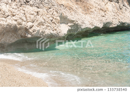 Close view of turquoise water meeting a small sandy beach beside a rocky cliff. Sunlight highlights the vivid coastal colors, capturing a peaceful and untouched seaside atmosphere. 133738540