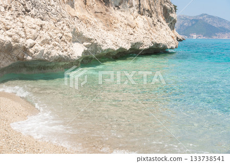 Shallow waves wash onto a light sandy beach beneath rocky cliffs. Clear turquoise water and bright sunlight create a serene coastal landscape with warm Mediterranean atmosphere. 133738541