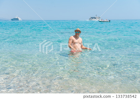 Person relaxing in shallow turquoise water with boats visible on the horizon. Clear sea, warm sunlight and calm conditions create a peaceful vacation scene ideal for travel themes Person relaxing in shallow turquoise water with boats visible on the horizon. Clear sea, warm sunlight and calm conditions create a peaceful vacation scene ideal for travel themes 133738542