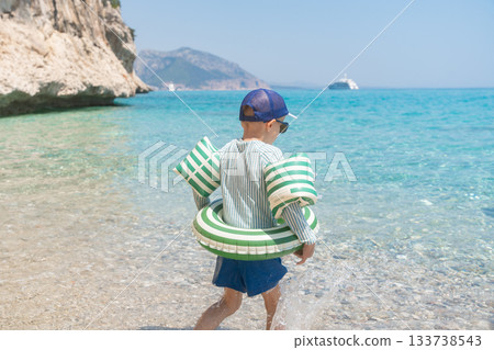 Child standing at the edge of a clear turquoise sea with rocky cliffs in the background. Bright sunlight highlights the shallow water and relaxed summer atmosphere of a quiet Mediterranean beach in 133738543