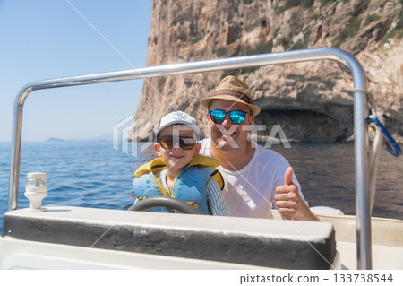 Adult and child enjoying a boat ride near rocky coastal cliffs. Bright sunlight, calm sea and smiling expressions create a warm vacation atmosphere ideal for travel and leisure themes 133738544