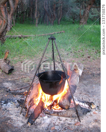 black pot hangs over a campfire in the forest on a tripod, vertical frame black pot hangs over a campfire in the forest on a tripod, vertical frame 133738876