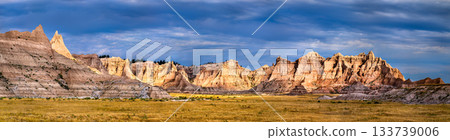 Panoramic view of eroded rock formations in Badlands National Park, South Dakota. Rugged buttes rise from the grassy prairie under a dramatic blue sky with dark clouds 133739006