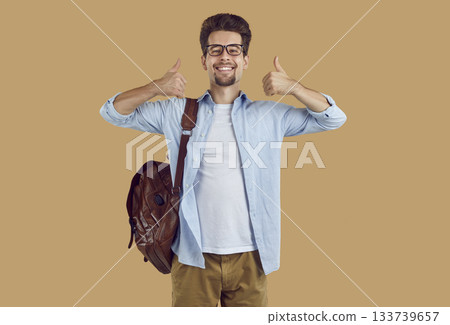 Happy university student guy with leather backpack showing thumbs up on beige background. 133739657