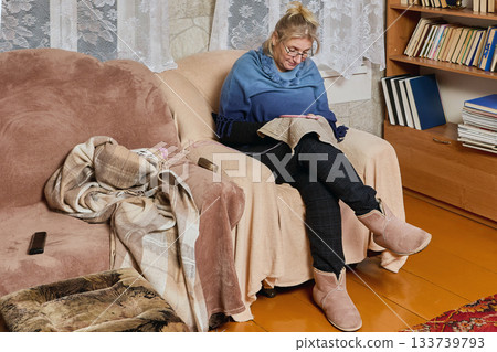 Process of embroidering within hoop caught middle aged woman in soft armchair in evening in rural house near book shelf. Process of embroidering within hoop caught middle aged woman in soft armchair in evening in rural house near book shelf. 133739793