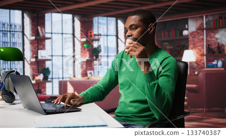 African american adult typing on a computer in his loft apartment, engaging in online freelancing work and multitasking efficiently. Flexibility of remote work in a cozy home office. Camera B. African american adult typing on a computer in his loft apartment, engaging in online freelancing work and multitasking efficiently. Flexibility of remote work in a cozy home office. Camera B. 133740387