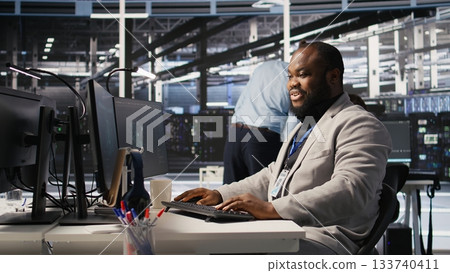 Joyous data center software developer delighted after solving all computer bugs. Smiling african american man in server hub pleased after finishing tasks on PC, overseeing server rigs, camera A 133740411