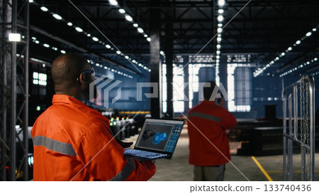 Engineer inspects turbine in industry plant and operating software on laptop to monitor heavy equipment performance. Warehouse features metal production tools and machinery. Engineer inspects turbine in industry plant and operating software on laptop to monitor heavy equipment performance. Warehouse features metal production tools and machinery. 133740436