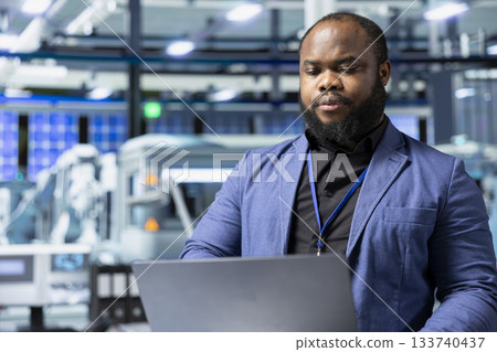 Solar energy black expert in a factory reviews data on laptop, inspecting manufacturing plant performance and symbolizing solar innovation. Industrial employee oversees production line. 133740437