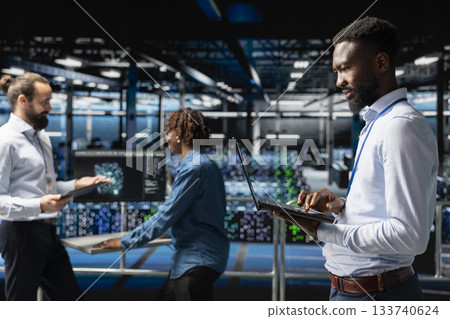 Black technician next to coworkers reviewing neural network code and LLM visualization dashboards. Server room IT specialist monitoring system performance with artificial intelligence tools. 133740624