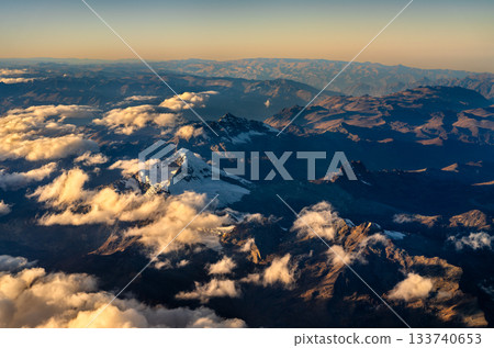 Aerial view of the Andes Mountains in Peru. Golden sunrise light illuminates the high peaks, while deep valleys and a sea of clouds are in shadow 133740653