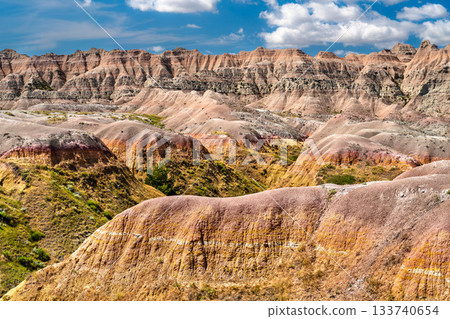 Colorful sedimentary rock layers in Badlands National Park, South Dakota. Eroded yellow and red hills rise under a blue sky with clouds Colorful sedimentary rock layers in Badlands National Park, South Dakota. Eroded yellow and red hills rise under a blue sky with clouds 133740654