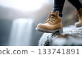 Close-up of rugged brown hiking boots on a rocky ledge, with a stunning waterfall cascading in the background, showcasing adventure and outdoor exploration 133741891