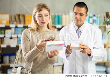 Young woman in pharmacy scans QR code on probiotic pills, man pharmacist in background 133742151