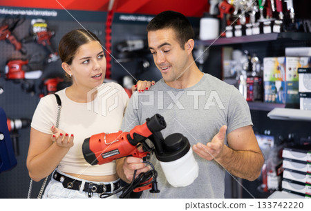 In store, spouses buyer doubts choice and closely examines electric paint-spraying machine spray gun In store, spouses buyer doubts choice and closely examines electric paint-spraying machine spray gun 133742220