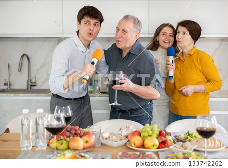 Happy family singing karaoke using microphone during family holiday, parents and adult children at festive table in kitchen Happy family singing karaoke using microphone during family holiday, parents and adult children at festive table in kitchen 133742262