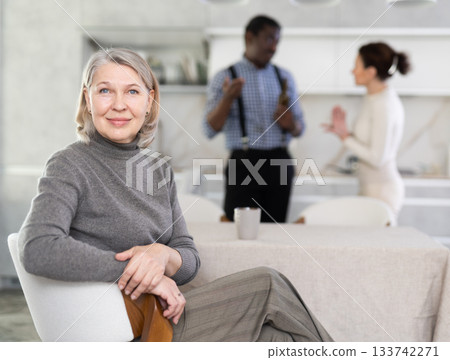 Smiling old woman sitting at the kitchen table with her back to couple quarreling together 133742271
