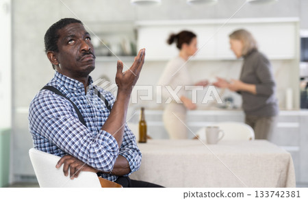 Middle-aged man sitting at the kitchen table with his back to women quarreling together Middle-aged man sitting at the kitchen table with his back to women quarreling together 133742381