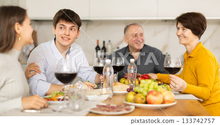 Smiling young man talking with family at festive table with wine and snacks at home 133742757