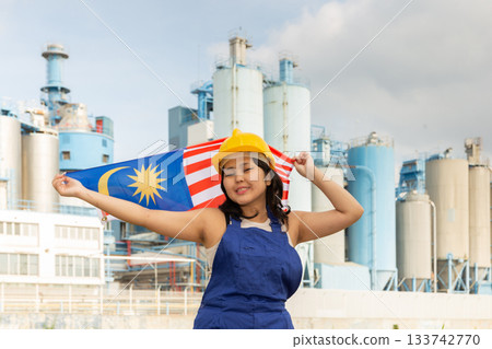 Portrait of positive girl in construction overalls with the Malaysia flag on the background of modern factory 133742770