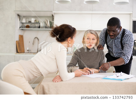 Old woman compiling testament while middle-aged man and woman competing with each other in the kitchen Old woman compiling testament while middle-aged man and woman competing with each other in the kitchen 133742793