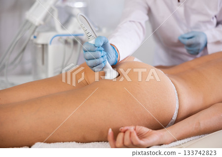 Cosmetologist performing high-frequency ultrasound procedure on buttocks of female client 133742823