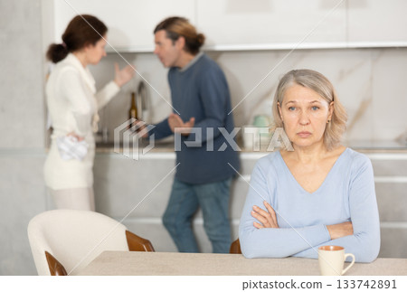 Frustrated senior woman is sitting in kitchen during quarrel between her son and his wife Frustrated senior woman is sitting in kitchen during quarrel between her son and his wife 133742891