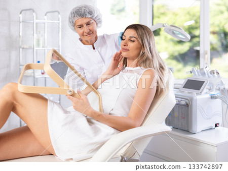 Young woman looking in mirror while mature cosmetologist showing results of facial procedure 133742897