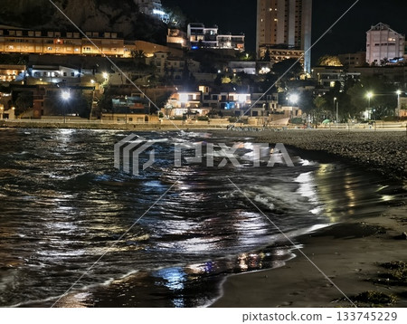 luminescent beach with cityscape, nighttime seafront illuminated by city lights and shimmering waves luminescent beach with cityscape, nighttime seafront illuminated by city lights and shimmering waves 133745229