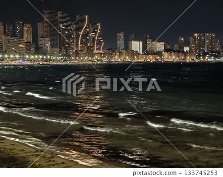 serene night seascape with city glow, nighttime ocean reflections with city illumination and gentle 133745253