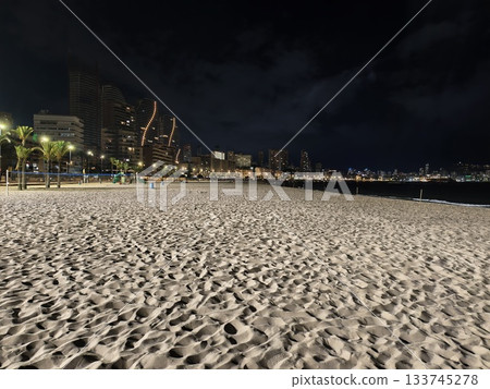 calm empty city beach at night with distant illuminated skyline and rippled sand foreground calm empty city beach at night with distant illuminated skyline and rippled sand foreground 133745278