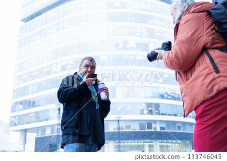 Female photographer in red jacket filming man with phone and coffee cup in snowy weather outside modern business center. High quality photo Female photographer in red jacket filming man with phone and coffee cup in snowy weather outside modern business center. High quality photo 133746045