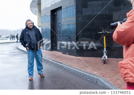 Female photographer in red jacket filming man in headphones listening to music in snowy rainy weather outside modern business center. High quality photo Female photographer in red jacket filming man in headphones listening to music in snowy rainy weather outside modern business center. High quality photo 133746051
