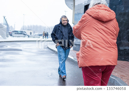 Female photographer in red jacket filming man in headphones listening to music in snowy rainy weather outside modern business center. High quality photo Female photographer in red jacket filming man in headphones listening to music in snowy rainy weather outside modern business center. High quality photo 133746058