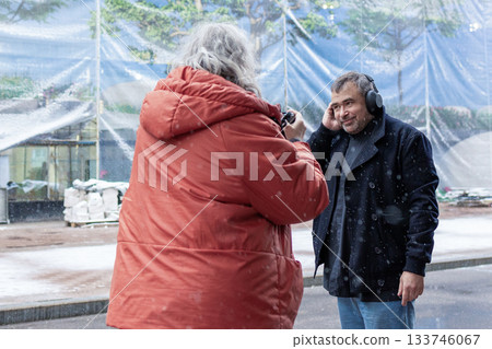 Female photographer in red jacket filming man in headphones listening to music in snowy rainy weather outside modern business center. High quality photo Female photographer in red jacket filming man in headphones listening to music in snowy rainy weather outside modern business center. High quality photo 133746067