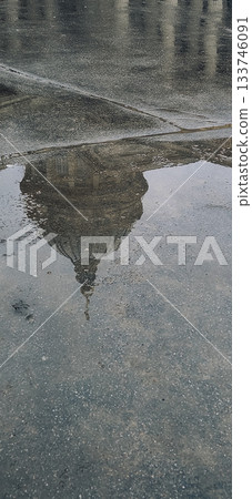 Rainy day city scene with architecture reflected in a puddle. People walk past, suggesting a quiet urban atmosphere. The photo captures a blend of weather and history.  133746091